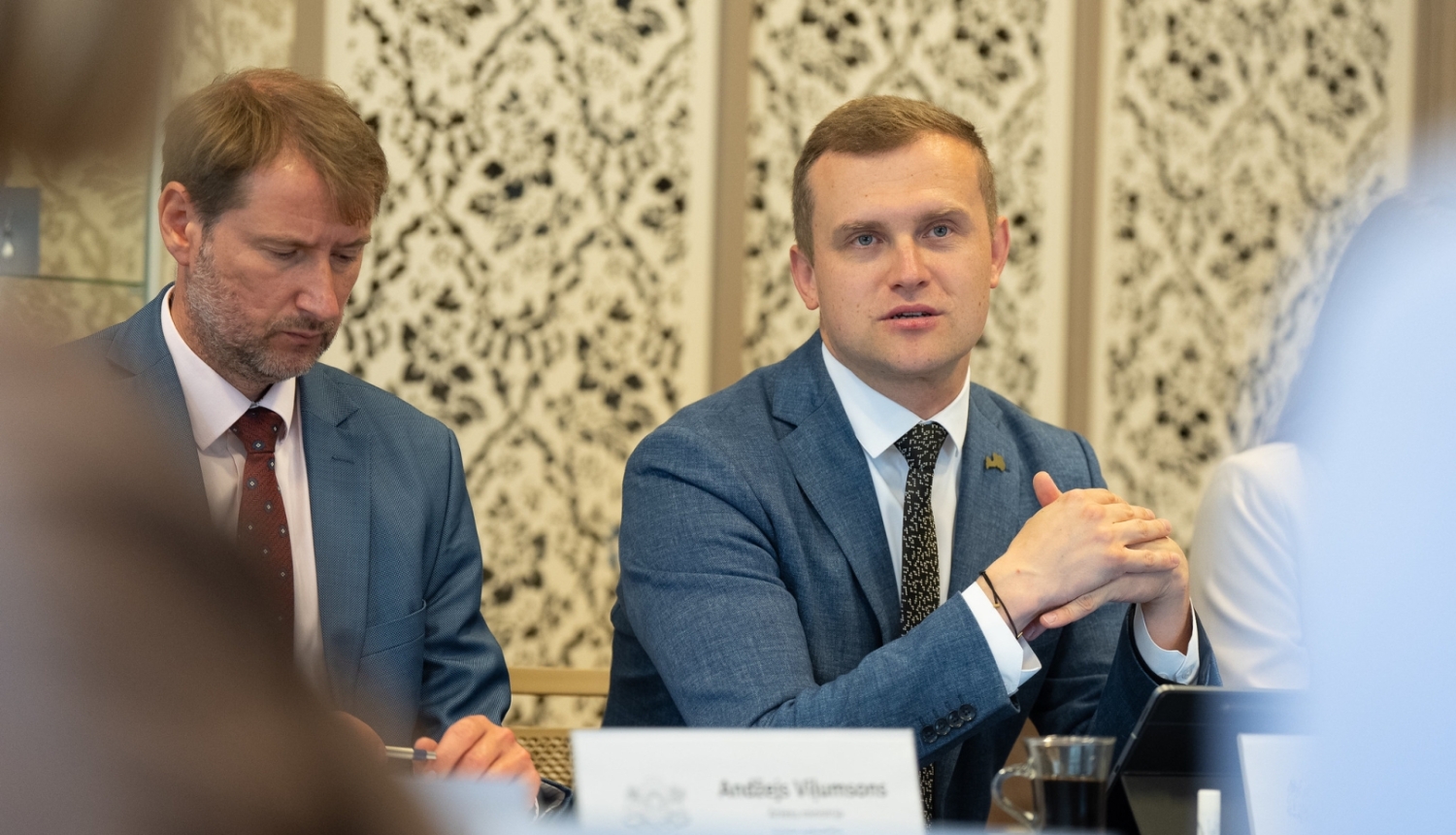 Parlamentārais sekretārs Artjoms Uršuļskis uzrunā klātesošos, viņam līdzās pie galda sēž valsts sekretārs Andžejs Viļumsons