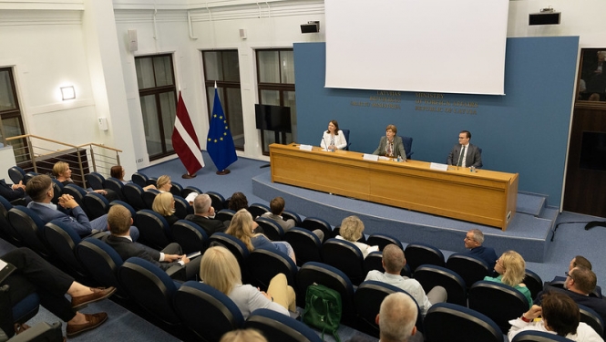 Ministre uzrunā pasākuma dalībniekus, kas sēž zālē