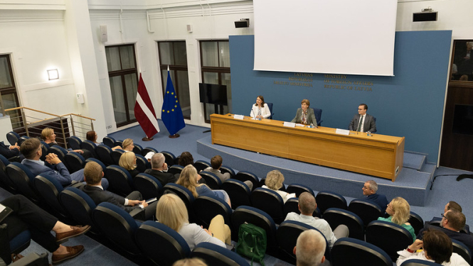 Ministre uzrunā pasākuma dalībniekus, kas sēž zālē