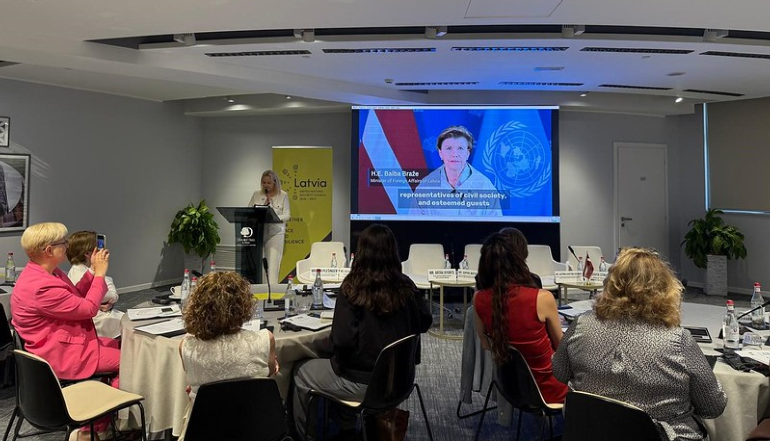 Dalībnieki pie apspriežu galda skatās Baibas Bražes uzrunu tiešsaistē